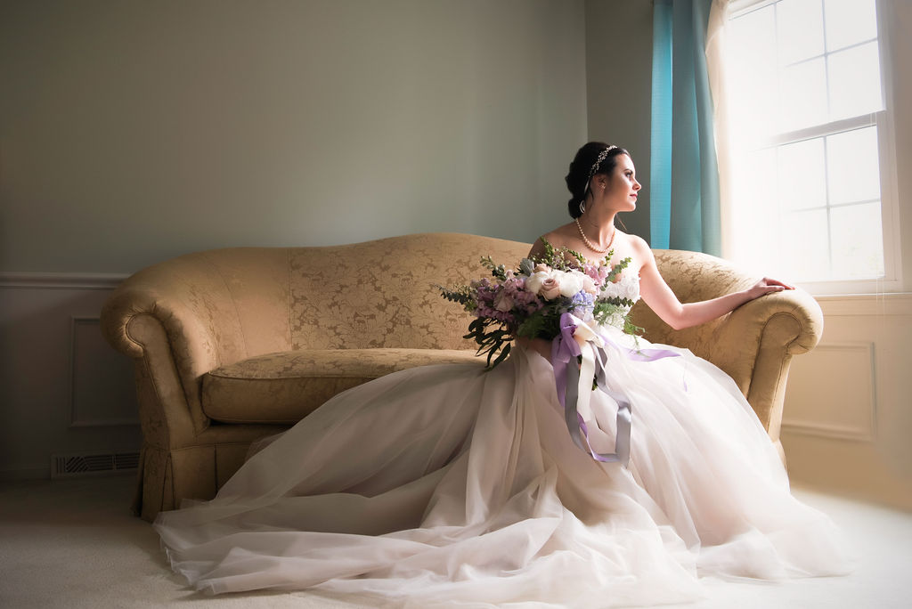 Bride poses elegantly on the couch and looks toward the window holding her beautiful bridal bouquet. 