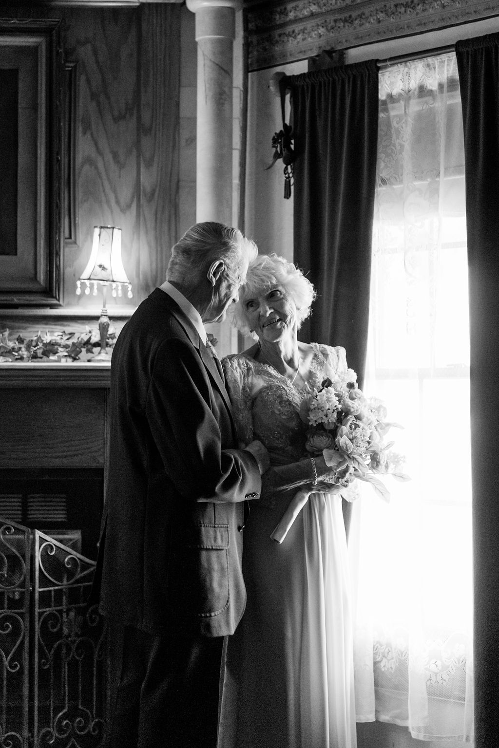 elderly bride and groom smile lovingly at each other in front of bright window