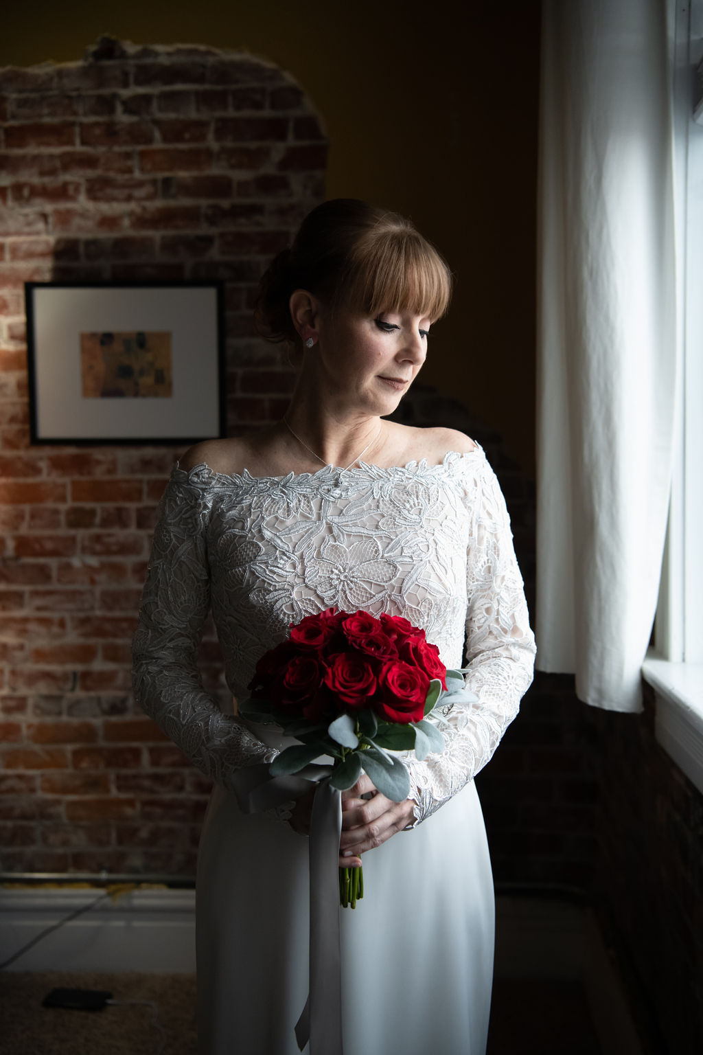 Bride glances down her shoulder towards the window holding her red bridal bouquet | choosing a getting ready space on your wedding day